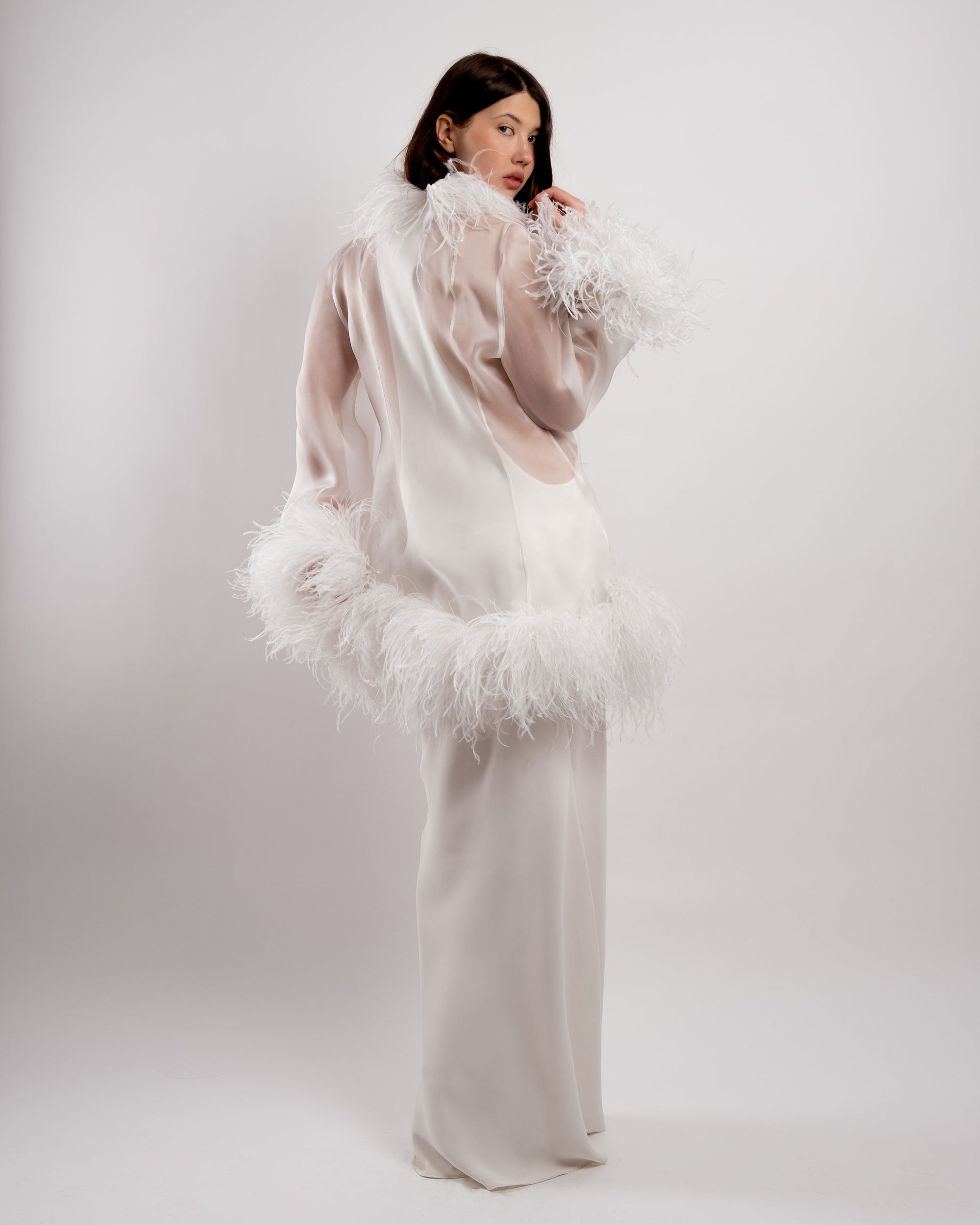 Woman wearing a white floor length gown with a ostrich feathered jacket against a white background
