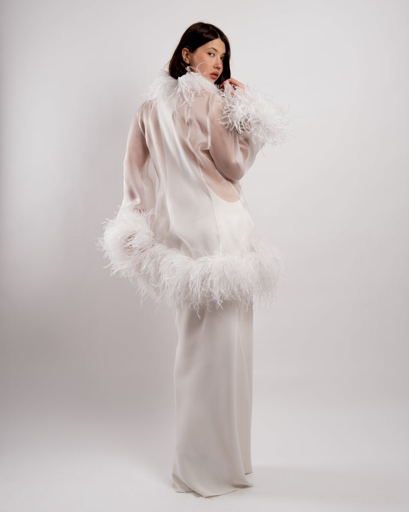 Woman wearing a white floor length gown with a ostrich feathered jacket against a white background