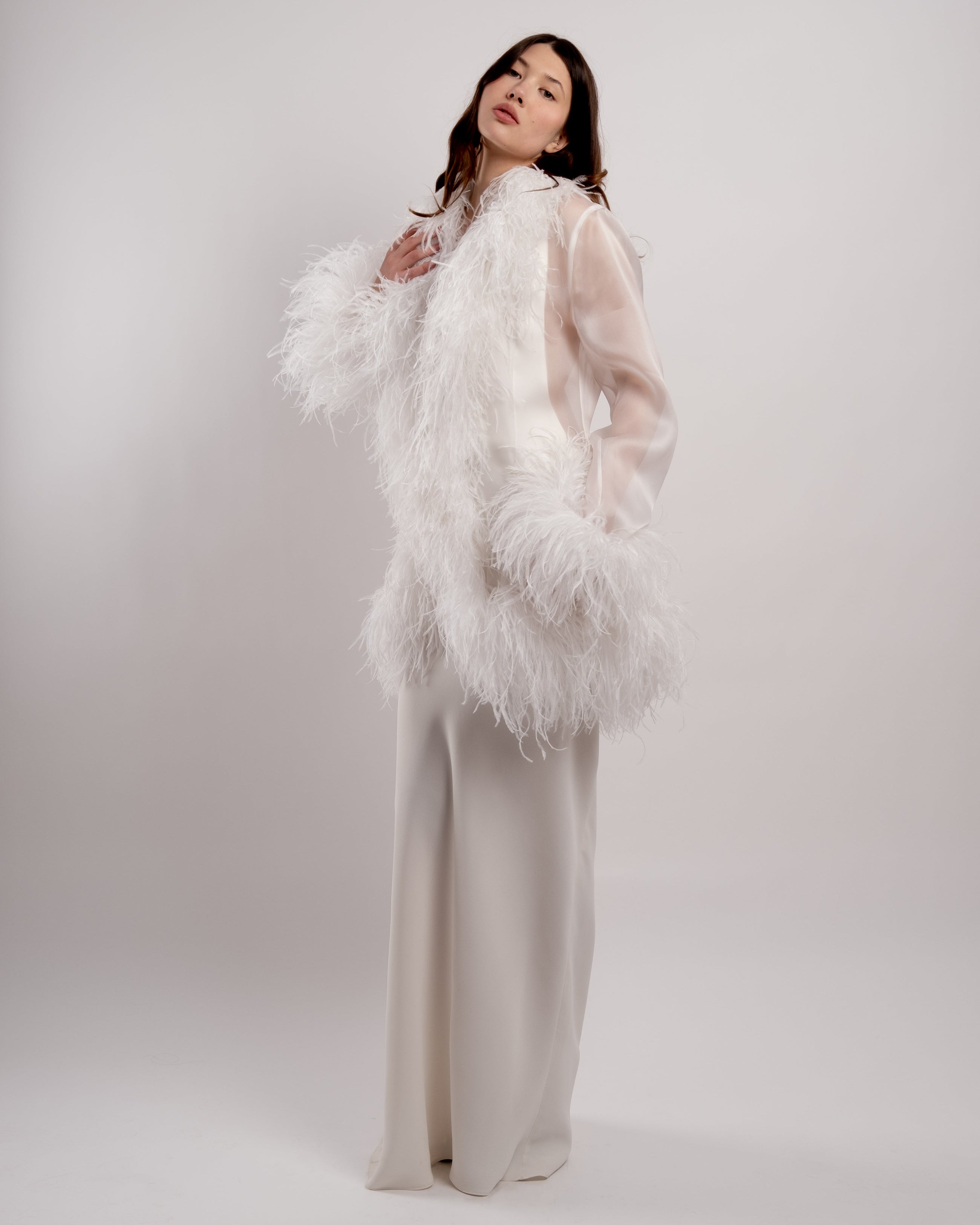 Woman wearing a white ostrich  feathered jacket and a white dress against a plain background