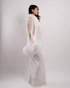 Woman wearing a long, elegant white dress with feathered details on a plain background