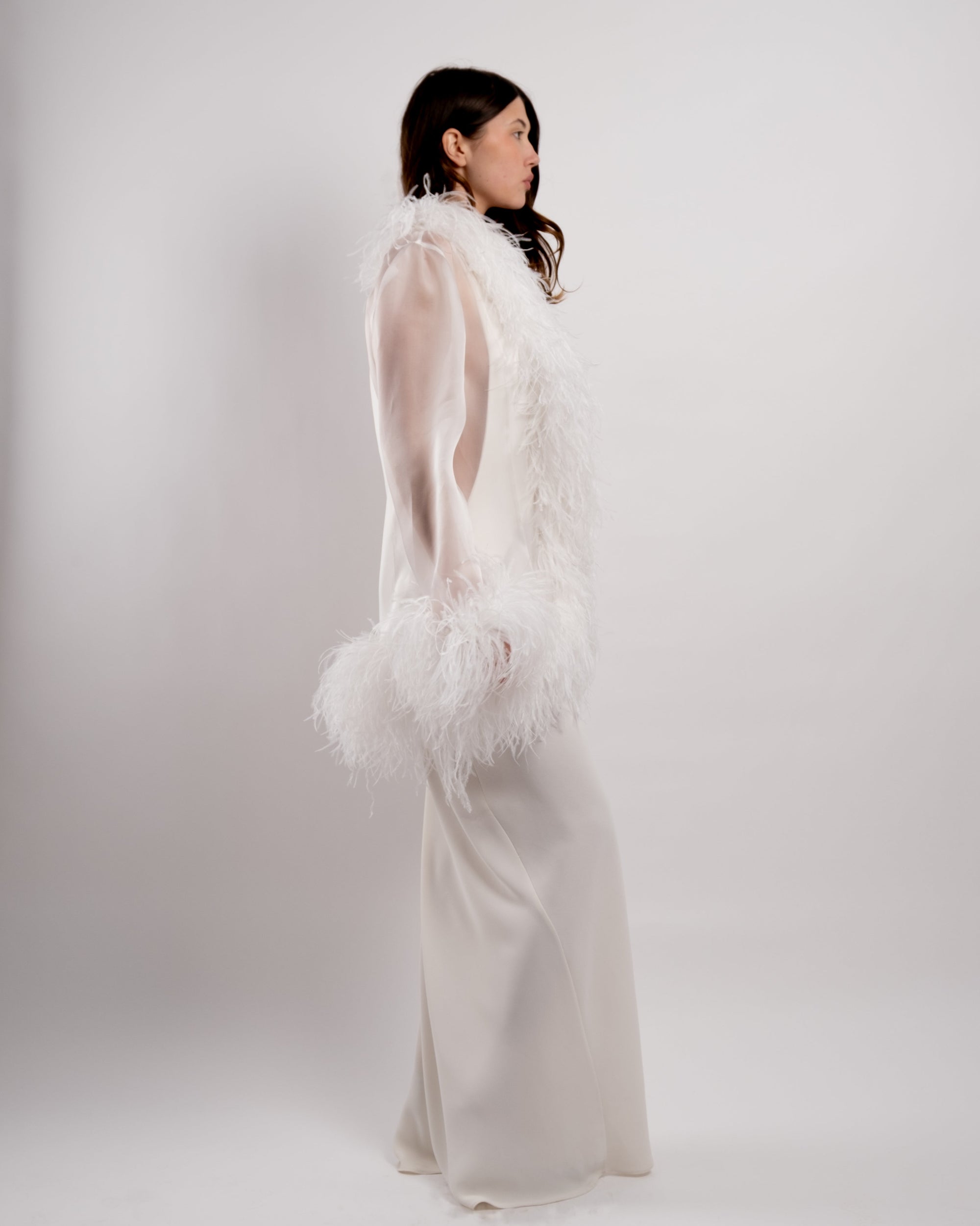 Woman wearing a long, elegant white dress with feathered details on a plain background