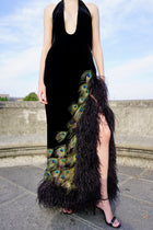 A model wearing a black velvet halter-neck maxi dress with a dramatic side slit covered in black feathers and decorated with peacock feather 